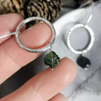 A close-up of a hand holding a hammered silver circle drop earring with a small carved green tourmaline leaf charm, with a second matching earring resting on a soft gray surface in the background.