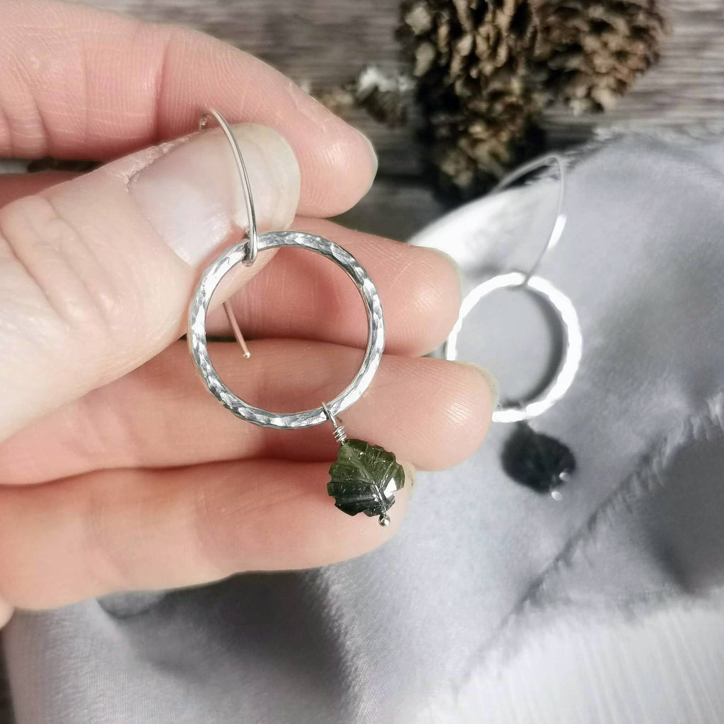 A close-up of a hand holding a textured silver circle drop earring with a small dark green gemstone dangle, with another earring resting on a gray fabric surface in the background.