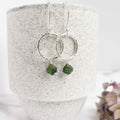 Silver circle drop earrings with green tourmaline leaf beads on a speckled grey ceramic surface