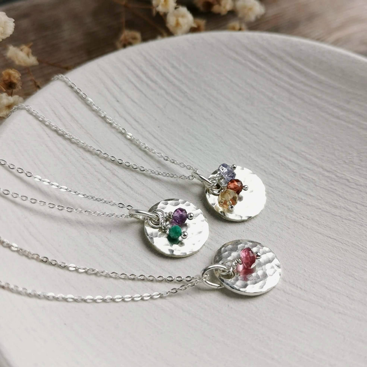 Three silver birthstone necklaces with hammered round pendants and small multicolor gemstone clusters for a variety of birth months from january to december, arranged on a light textured surface with dried flowers in the background.