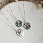 Three silver birthstone necklaces with hammered round disc pendants and small colorful gemstone charms for may birth months from january to december, arranged on a white textured surface with dried flowers in the background.