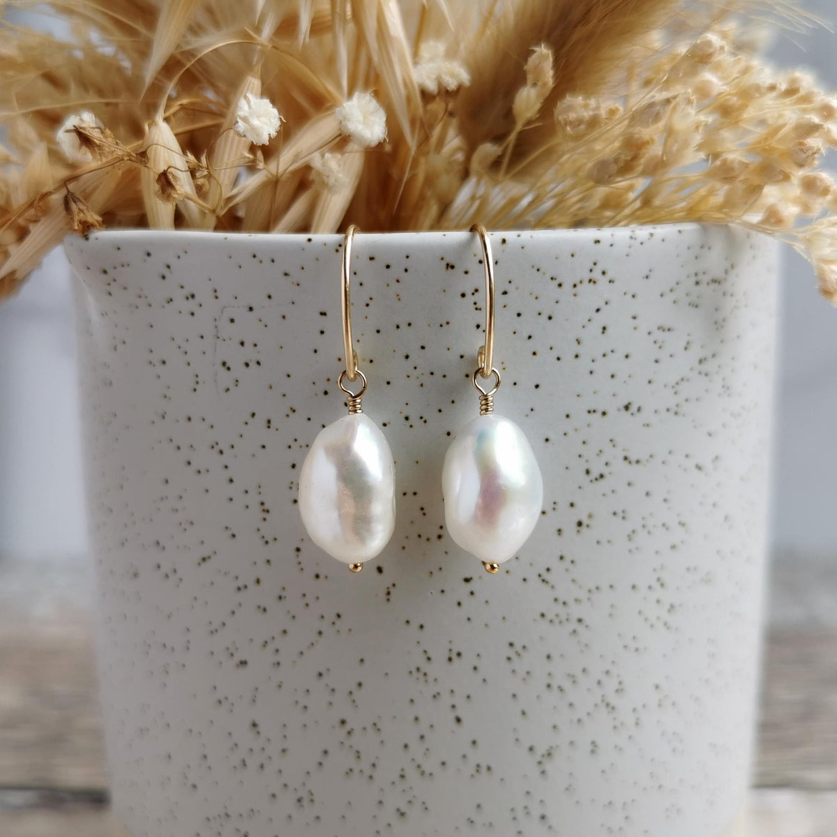 A pair of 14k gold-filled drop earrings with large, irregular ivory white baroque pearls, hanging from a speckled white ceramic pot filled with dried grasses.