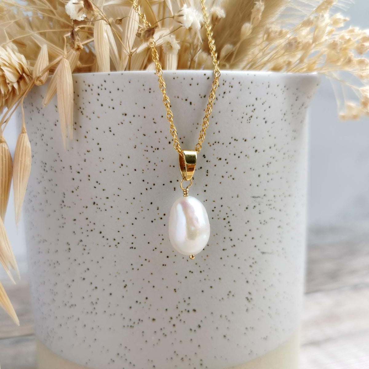A 14k gold-filled chain necklace with an ivory baroque pearl pendant, displayed hanging from a speckled white ceramic pot filled with dried grasses.