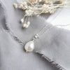 A silver or 14k gold-filled pendant necklace with a single baroque pearl centerpiece, presented on a grey fabric surface with dried flowers.