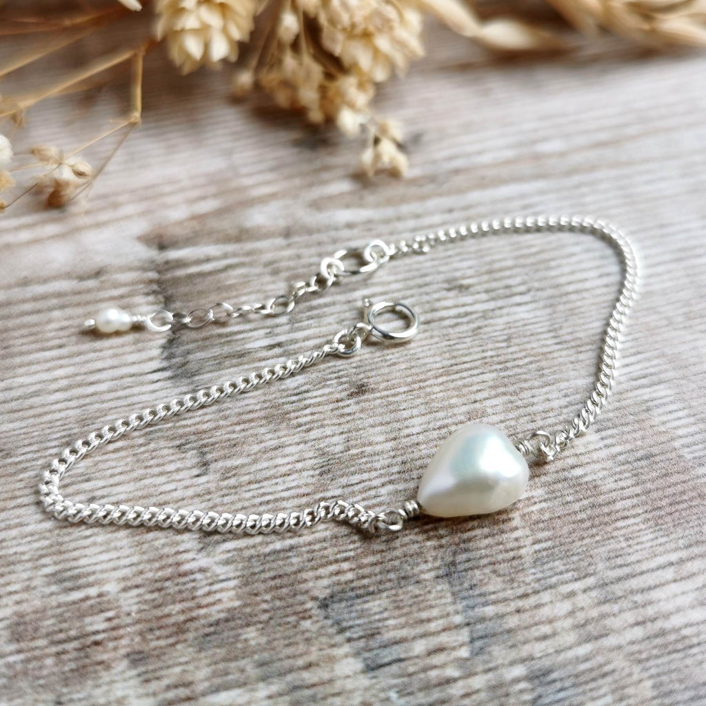 A delicate silver chain bracelet featuring a single irregular ivory white freshwater pearl, displayed on a rustic wooden surface with dried flowers in the background.