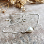 A delicate silver chain bracelet featuring a single irregular ivory white freshwater pearl, displayed on a rustic wooden surface with dried flowers in the background.