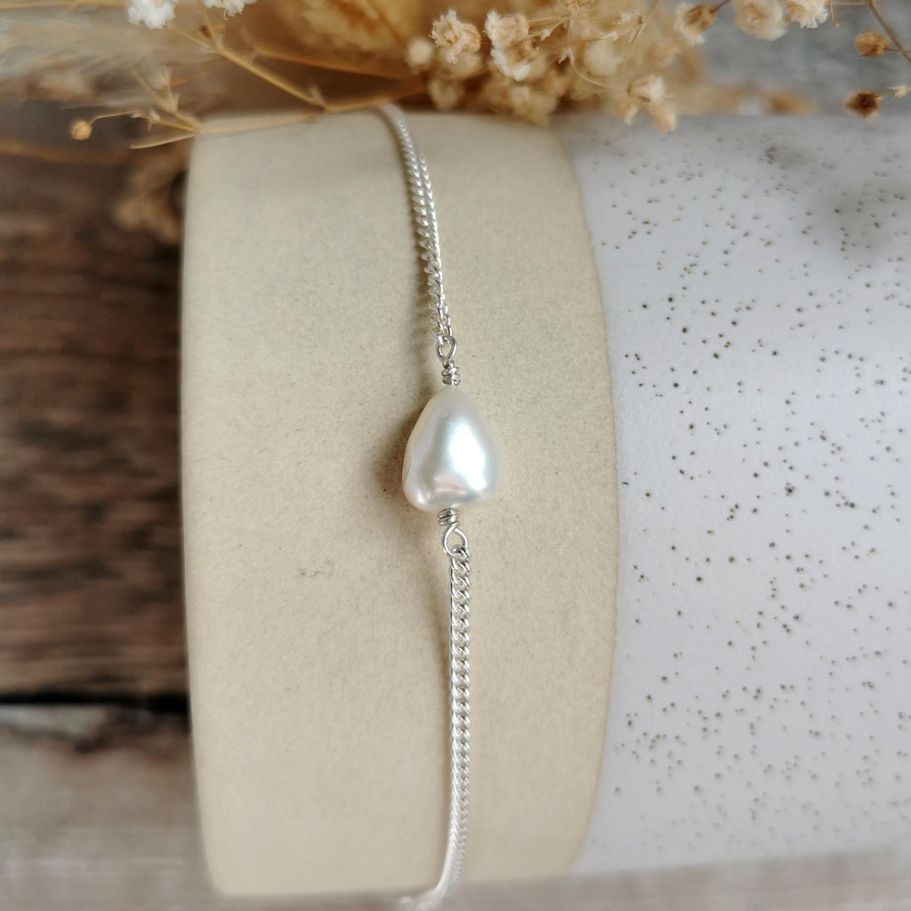 A delicate silver chain bracelet featuring a single teardrop-shaped white freshwater baroque pearl, displayed on a beige cylindrical stand with dried flowers in the background.