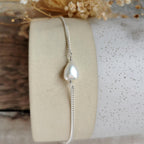 A delicate silver chain bracelet featuring a single teardrop-shaped white freshwater baroque pearl, displayed on a beige cylindrical stand with dried flowers in the background.