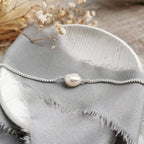 A delicate silver chain bracelet featuring a single irregular white freshwater baroque pearl, styled on a textured gray fabric with dried flowers.