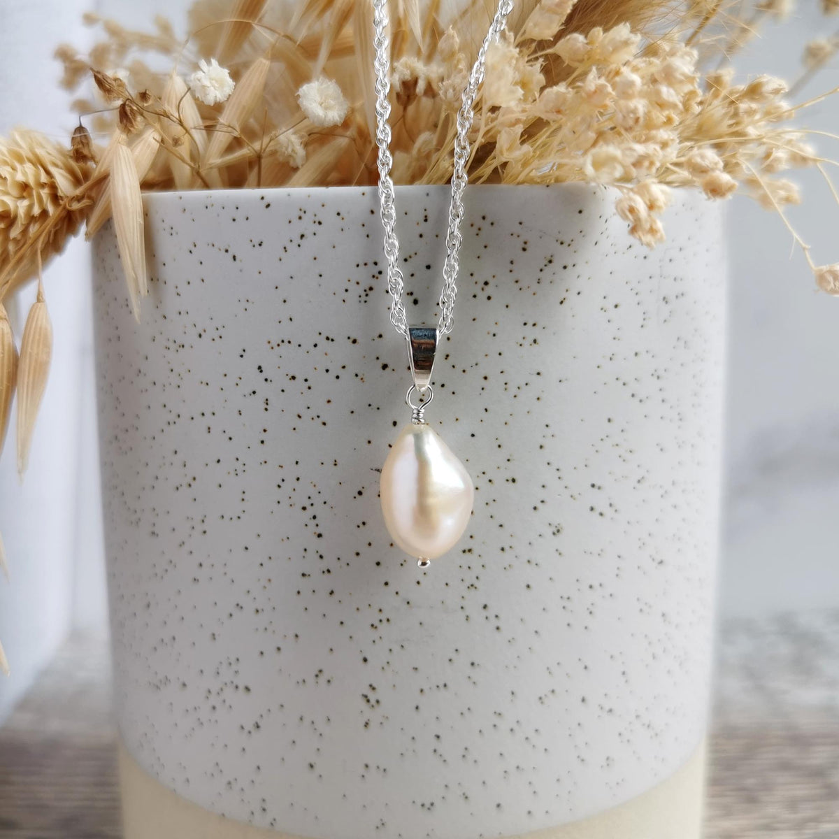 A silver chain necklace with a large, irregular ivory baroque pearl pendant, hanging in front of a speckled white ceramic pot filled with dried grasses.
