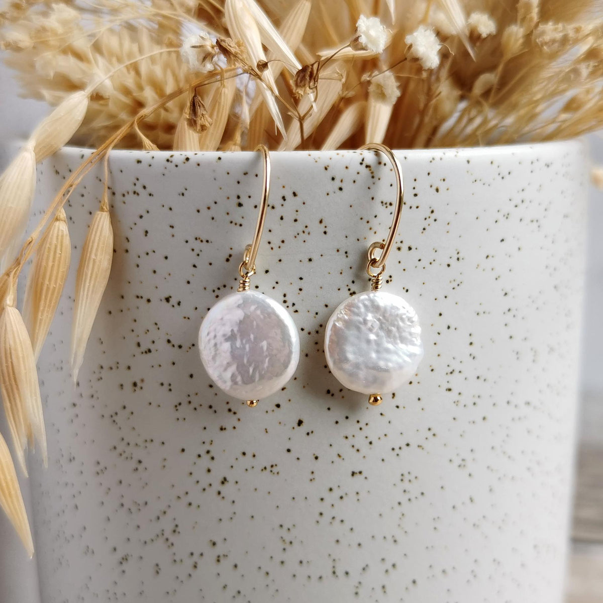 A pair of handmade 14k gold-filled drop earrings with textured, irregular ivory coin pearls, displayed on a speckled white ceramic cup with dried grasses.