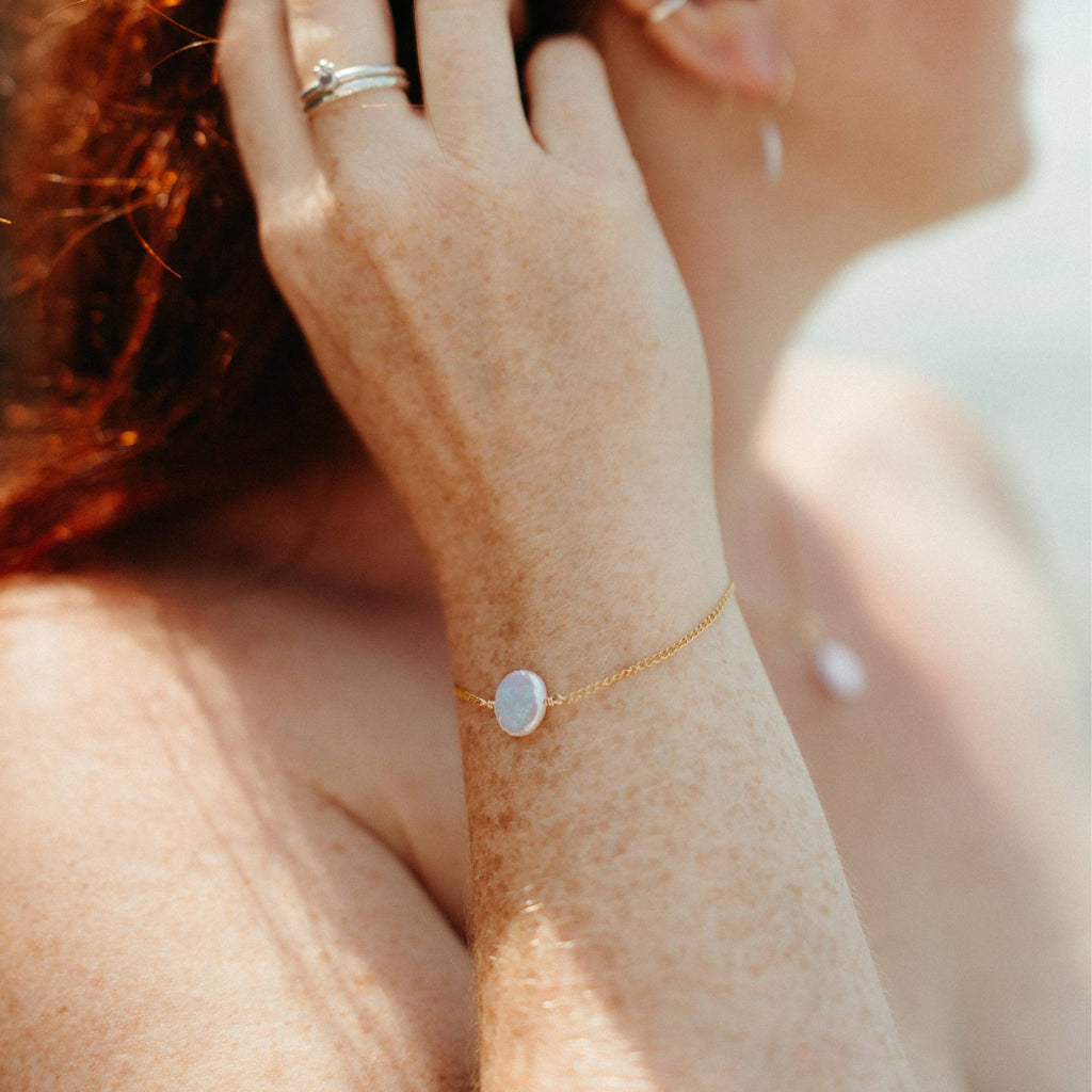 A close-up lifestyle shot of a woman's wrist wearing a delicate gold chain bracelet with a smooth, round ivory pearl. The image is shot in warm, bright sunlight, highlighting the skin texture and the jewelry.