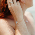 A close-up lifestyle shot of a woman's wrist wearing a delicate gold chain bracelet with a smooth, round ivory pearl. The image is shot in warm, bright sunlight, highlighting the skin texture and the jewelry.
