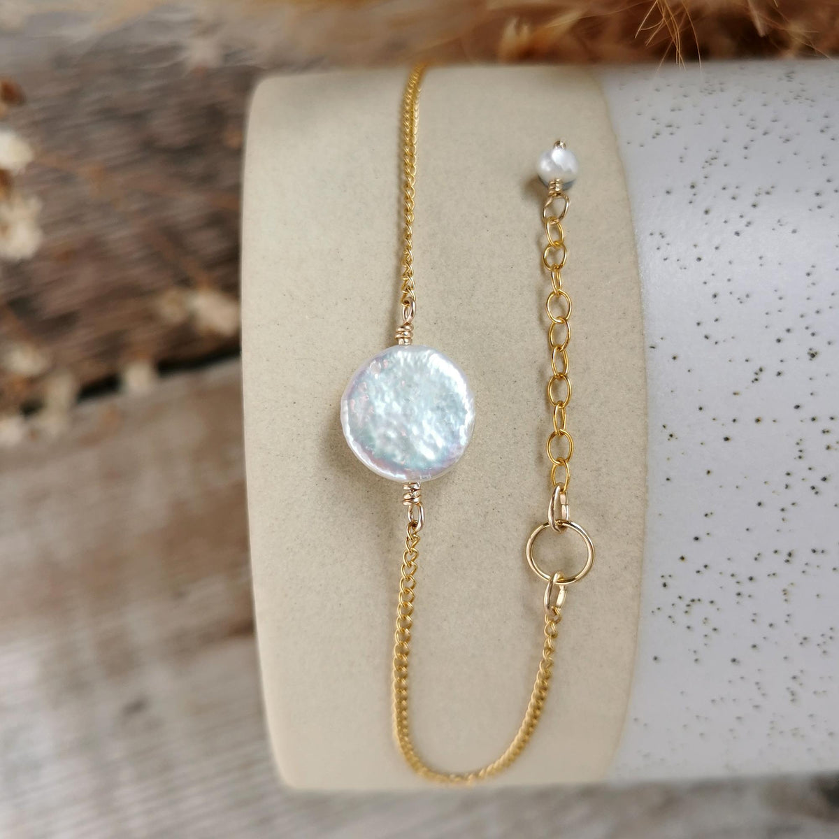 A delicate gold chain bracelet with a small, textured ivory white coin pearl and a smaller matching pearl on an adjustable chain, displayed on a beige jewelry stand.
