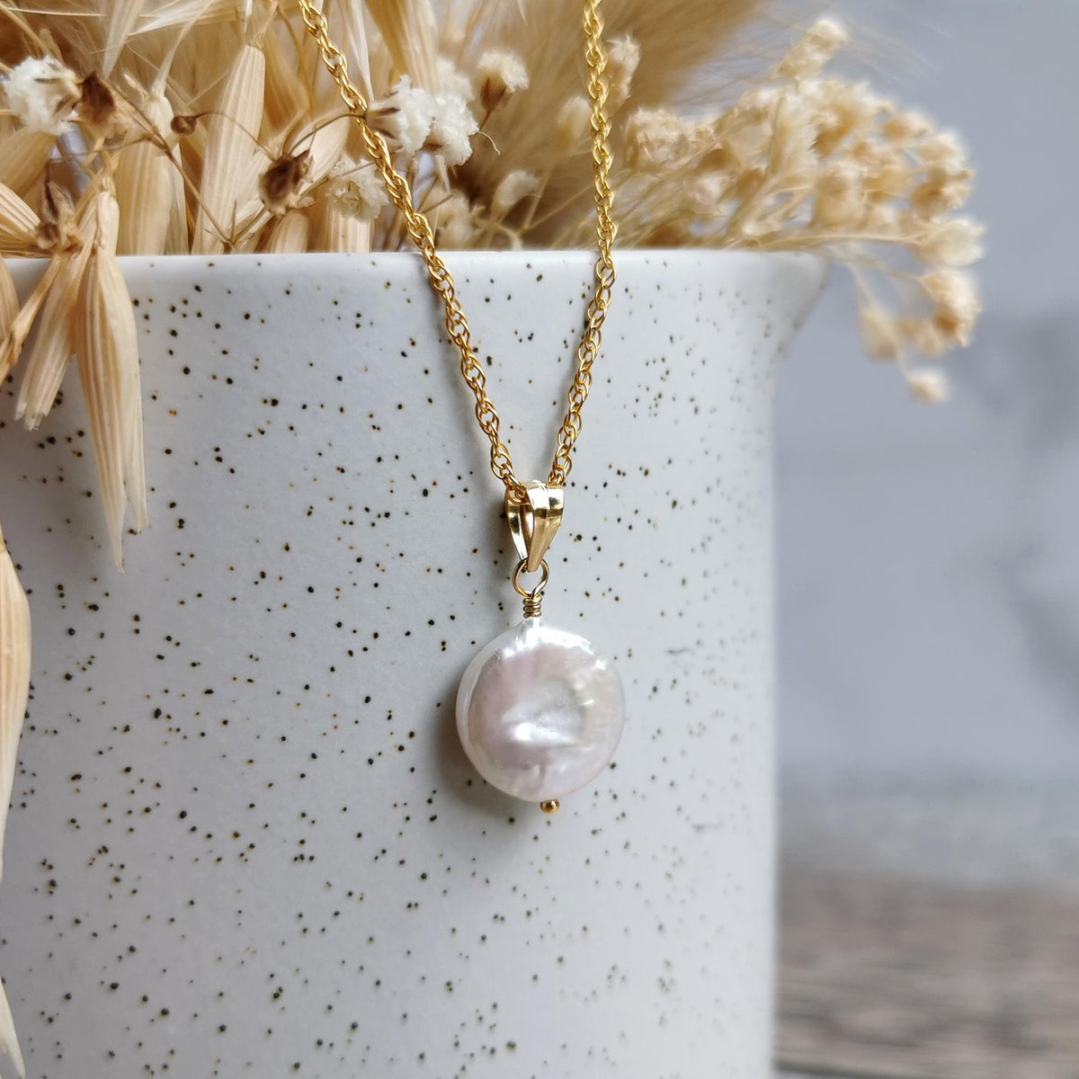 A gold chain necklace with a small, irregular white coin pearl pendant, styled draped over a speckled white ceramic vase with dried grass.