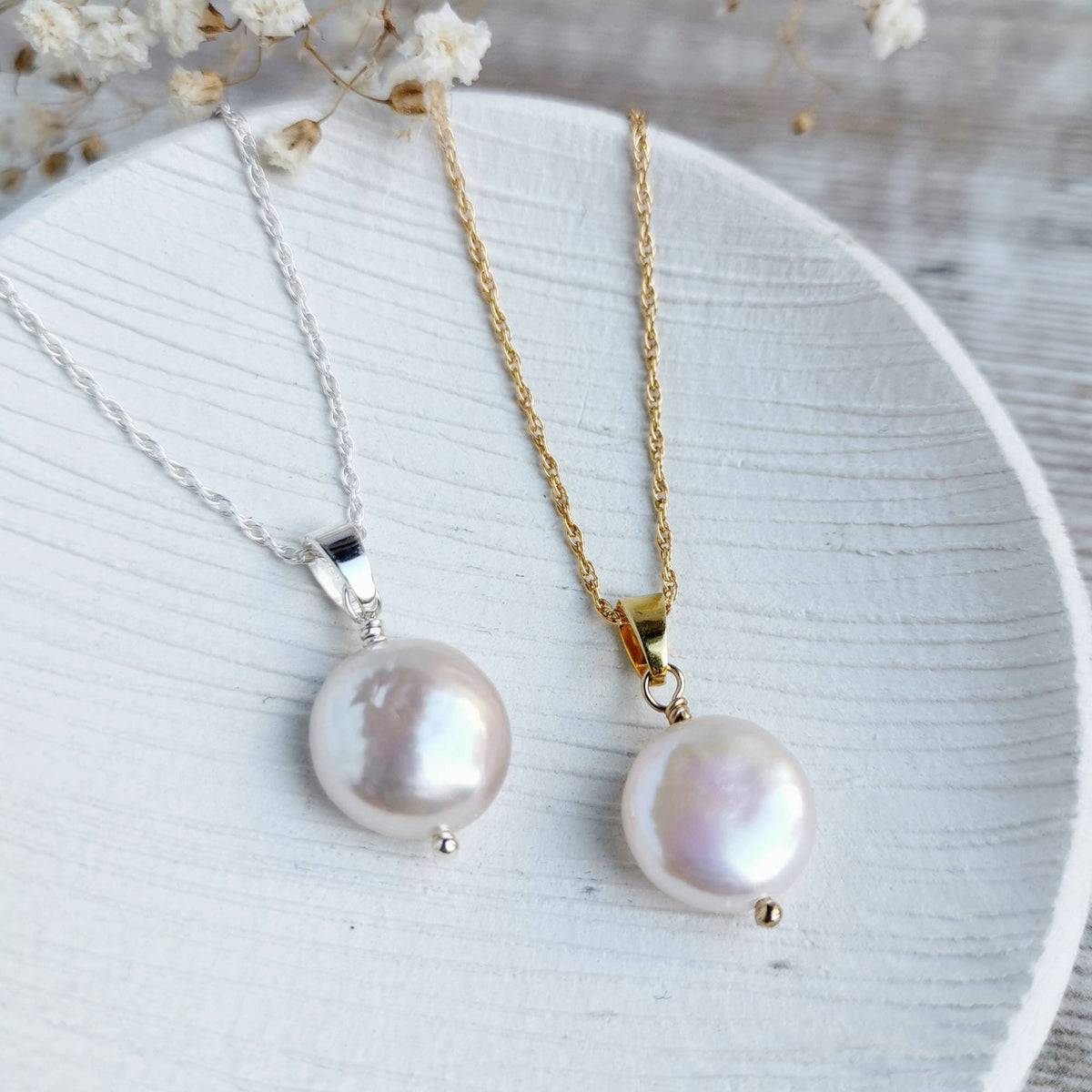 Two freshwater coin pearl necklaces, one with a silver chain and bail and one with a gold chain and bail, displayed on a white textured ceramic plate with dried flowers.