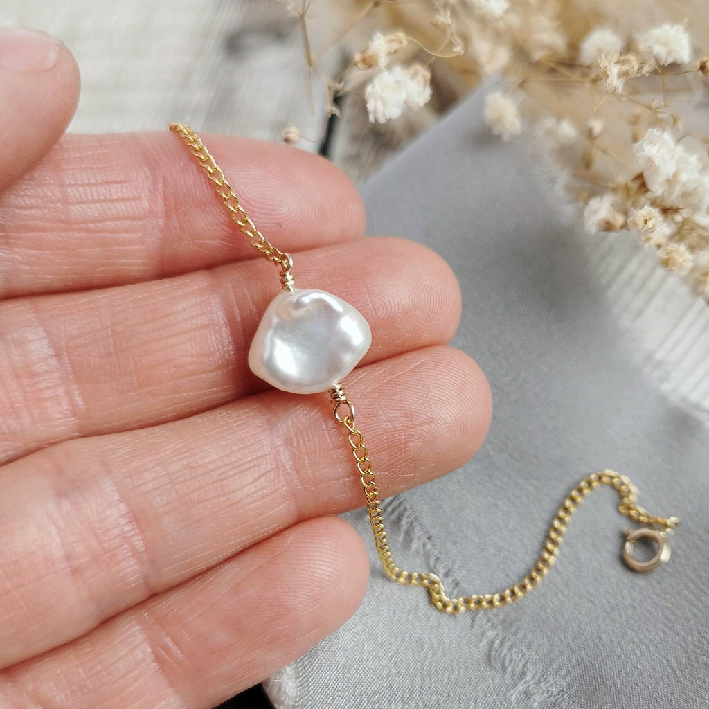 Gold chain bracelet with an ivory keishi pearl held on fingers on a neutral background