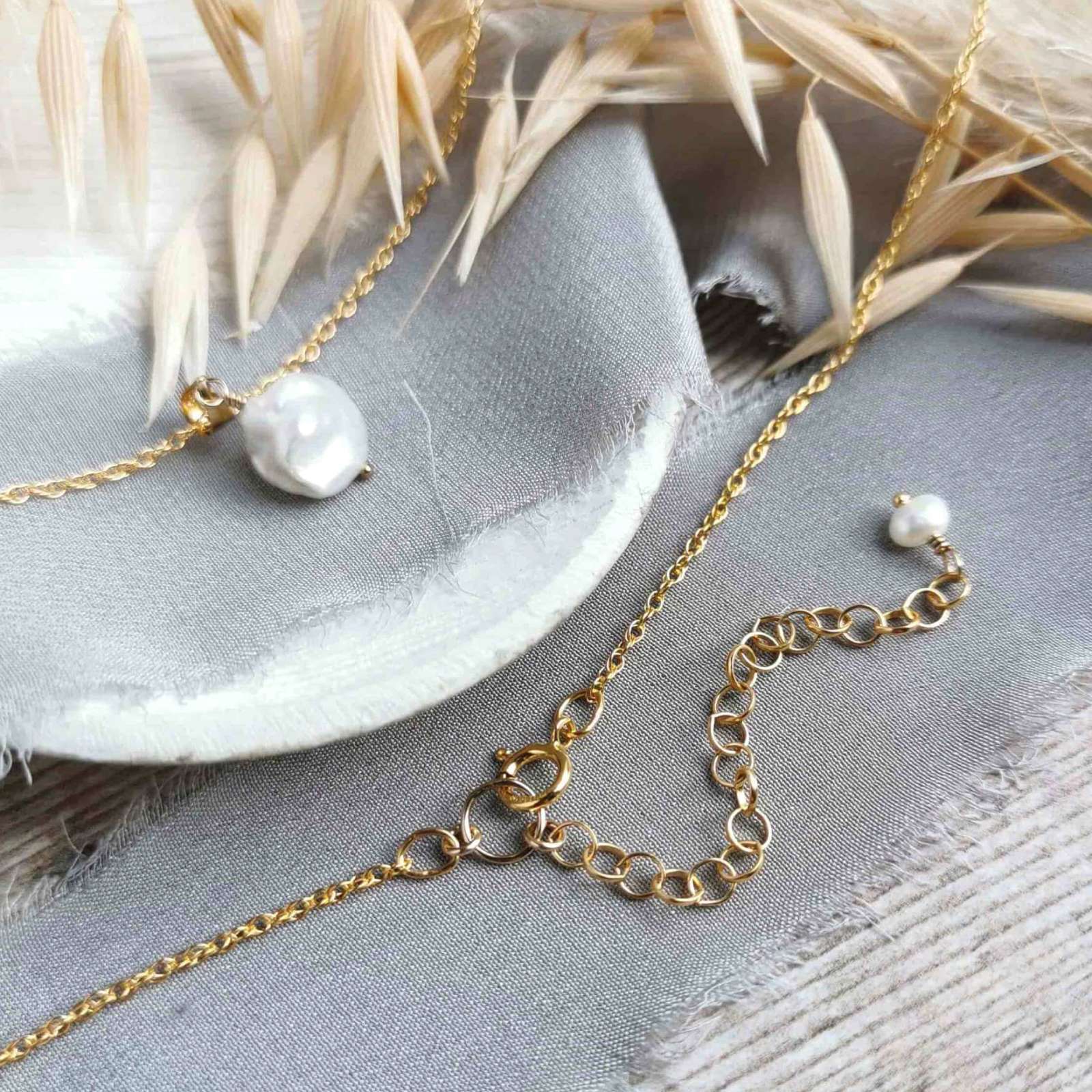 Gold and pearl necklace with extender chain on a light grey surface with dried leaves.