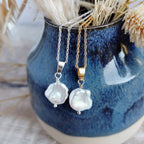 Two pearl necklaces on gold and silver chains in front of a blue ceramic vase with dried flowers.