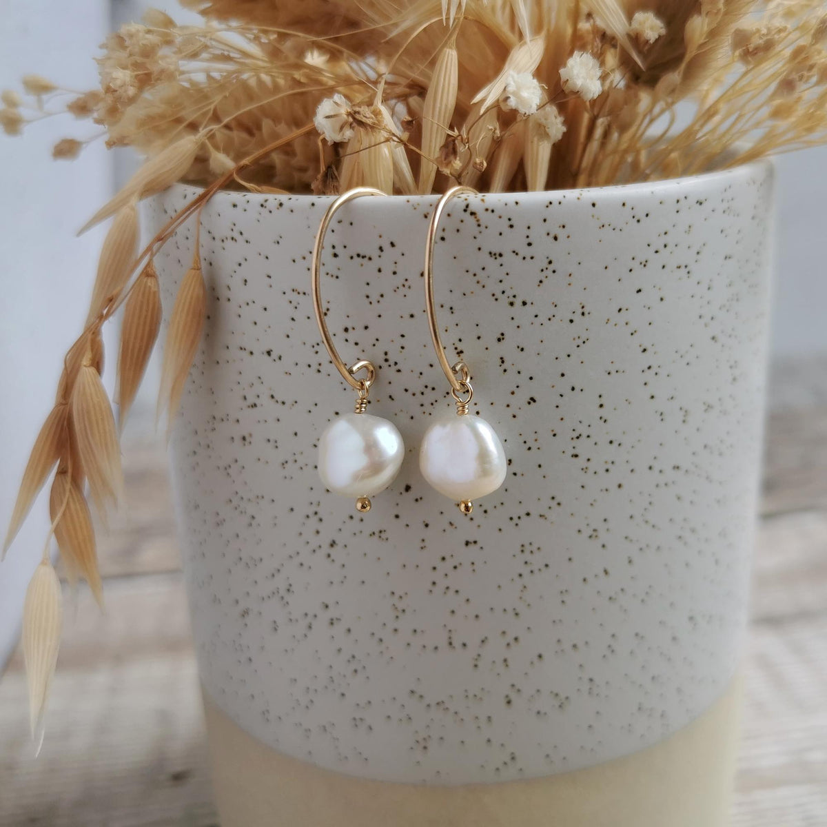 A pair of 14k gold-filled earrings with irregular ivory white freshwater keshi pearls hanging from a speckled ceramic pot filled with dried grasses.