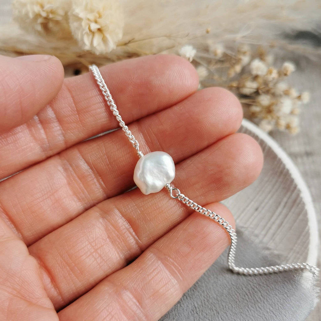 A close-up of a hand holding a delicate handmade silver chain bracelet with a single petal shaped ivory white keshi pearl, with dried flowers in the background.