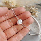 A close-up of a hand holding a delicate handmade silver chain bracelet with a single petal shaped ivory white keshi pearl, with dried flowers in the background.