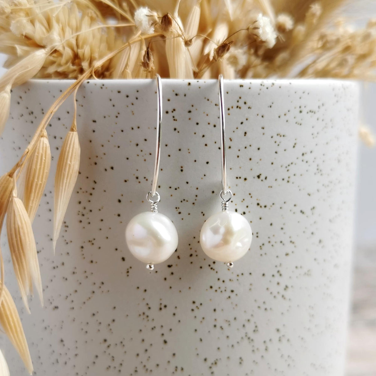 A pair of silver drop earrings with lustrous plump ivory white keshi pearls, hanging from a speckled white ceramic pot with dried grasses behind them.