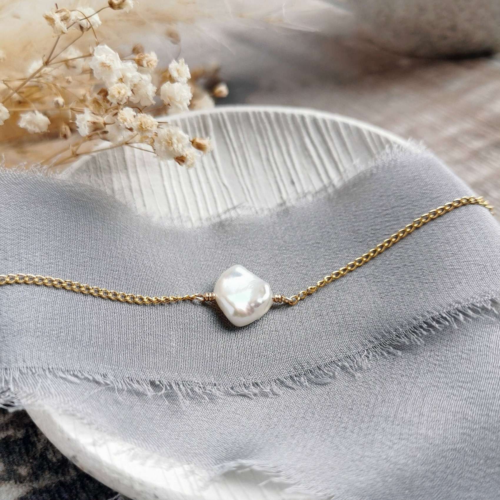 Gold chain bracelet with a unique flat pearl on a light grey fabric background