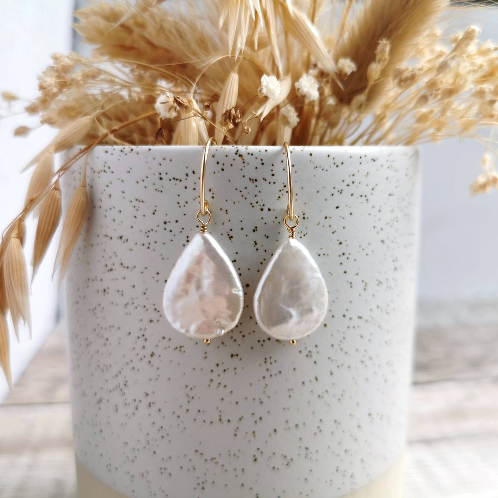 A pair of teardrop-shaped drop earrings made from iridescent ivory white pearls with gold earwires, displayed on a speckled white ceramic pot filled with dried grasses.