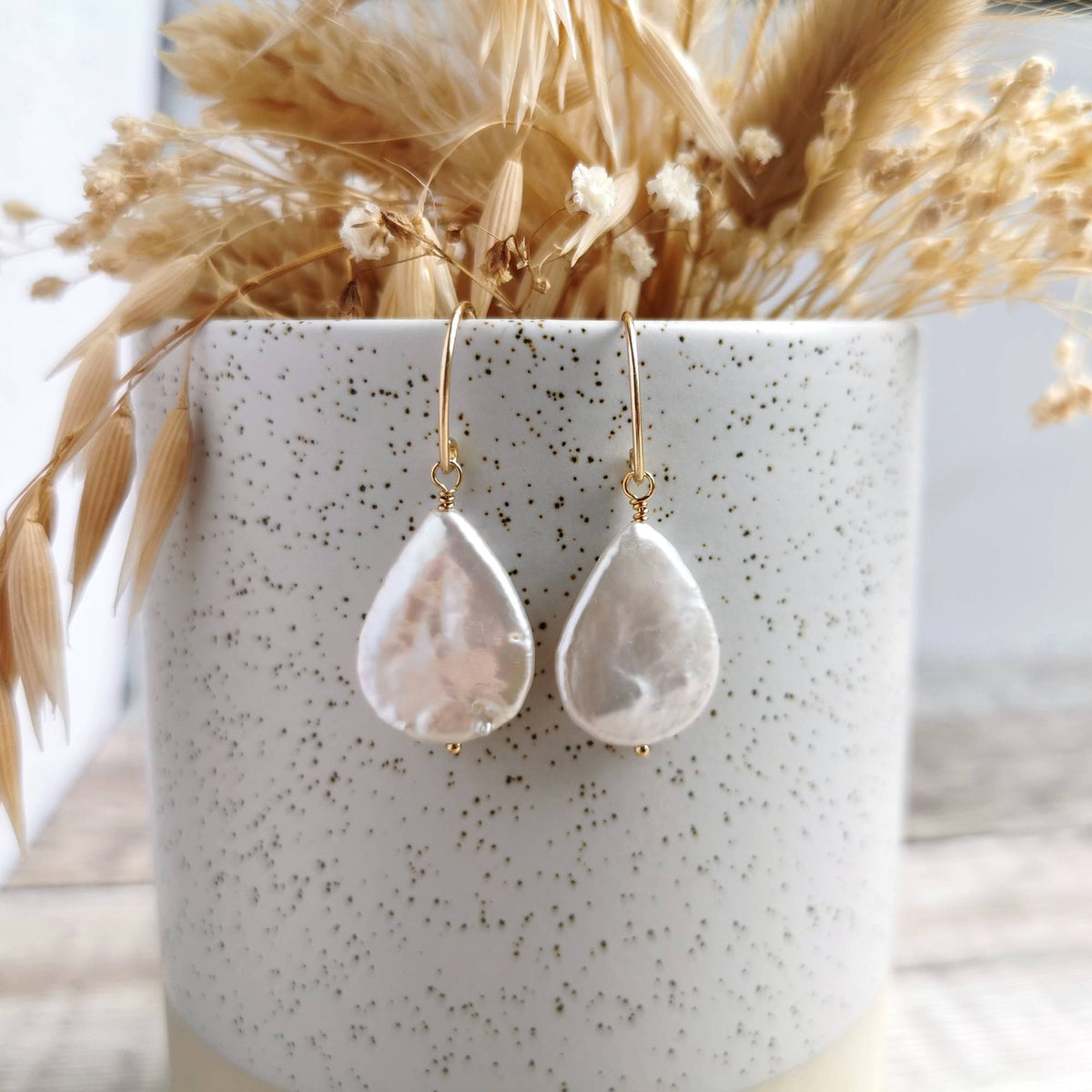 A pair of teardrop-shaped drop earrings made from iridescent ivory white pearls with gold earwires, displayed on a speckled white ceramic pot filled with dried grasses.