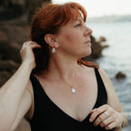 A woman with red hair wearing a black swimsuit and matching teardrop pearl earrings and necklace, posing outdoors near the water with rocks on the coast in Cornwall.