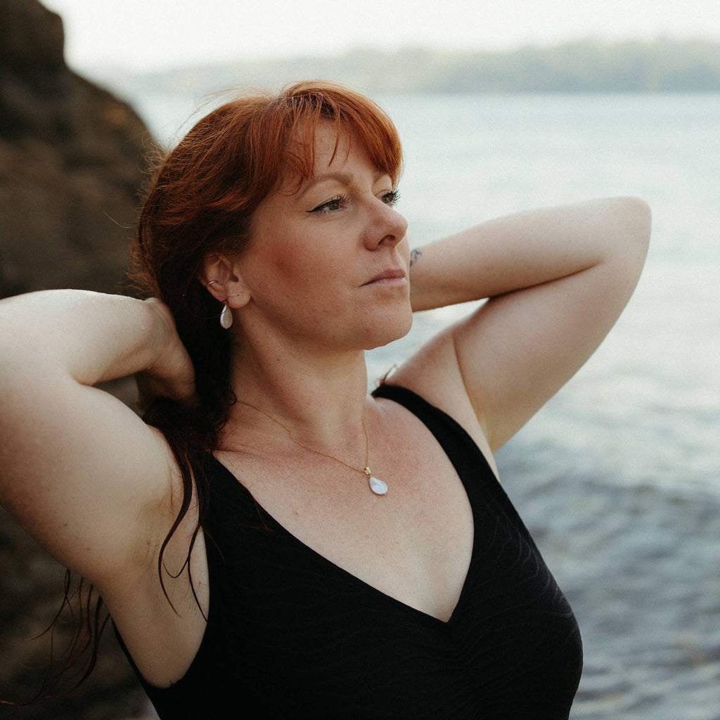 A woman with red hair posing outdoors by the water, wearing a black swimsuit and an ivory white teardrop pearl pendant necklace and matching earrings.