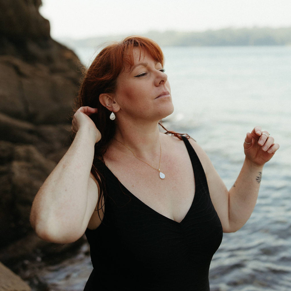 A woman with red hair stands by the water wearing a black swimsuit with teardrop pearl and 14k gold-filled necklace and earrings set, with rocks and a blurred cornish shoreline in the background.