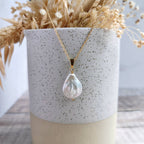 A gold chain necklace with an ivory teardrop-shaped flat baroque pearl pendant, displayed on a speckled ceramic vase with dried flowers.