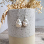A pair of teardrop-shaped pearl drop earrings with textured off-white pearls on silver hooks, hanging from a speckled ceramic vase filled with dried grasses.