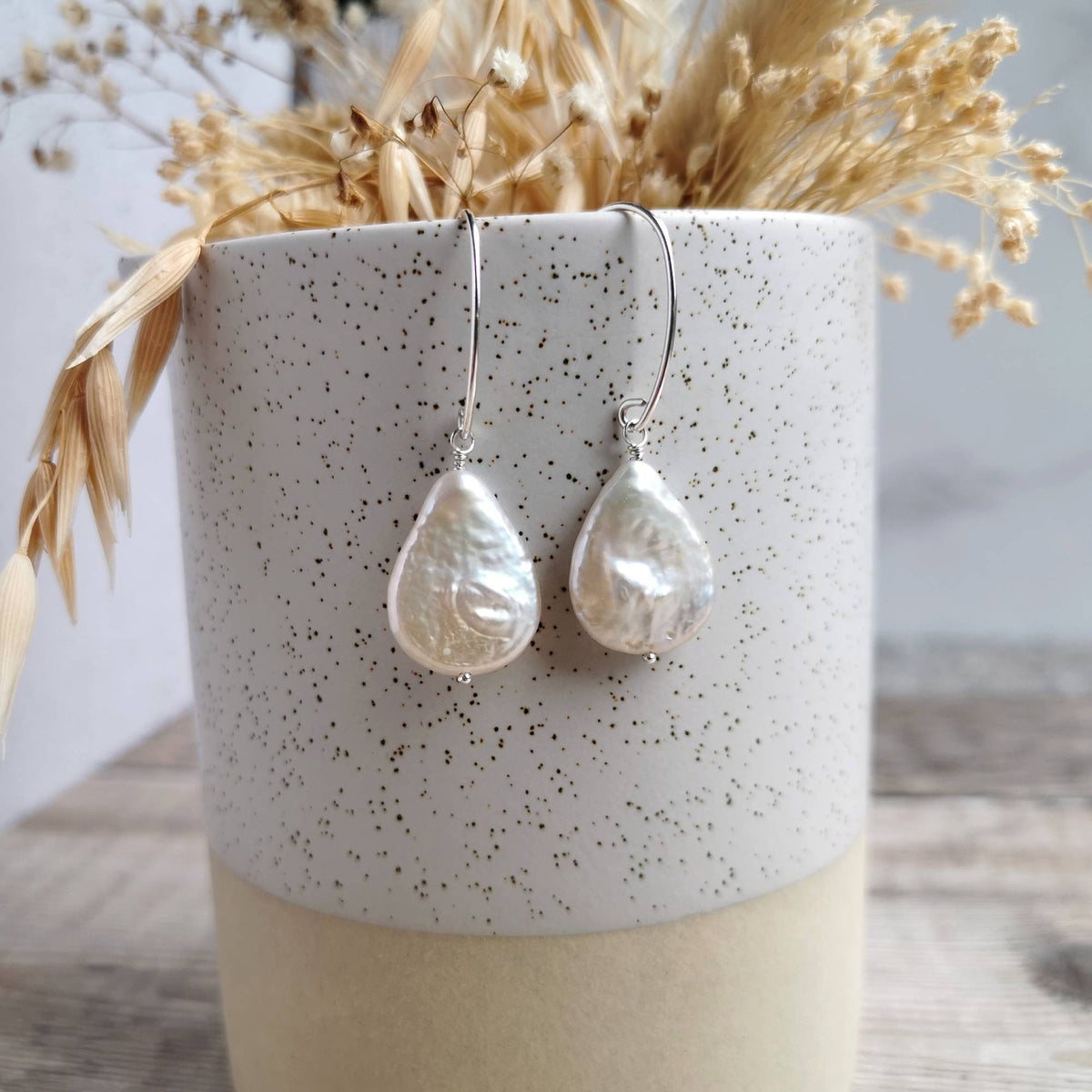 A pair of teardrop-shaped pearl drop earrings with textured off-white pearls on silver hooks, hanging from a speckled ceramic vase filled with dried grasses.