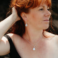 A woman with red hair wearing a silver and freshwater teardrop pearl necklace and matching earrings, and a delicate silver bracelet, shown in a close-up profile shot.
