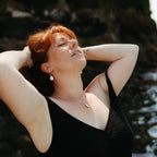 A woman with red hair wearing a black swimsuit and silver with teardrop pearl jewellery, posing outdoors against a dark rocky coastal background near falmouth, cornwall.