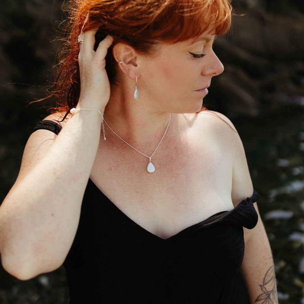 A woman with red hair wearing a delicate silver necklace with a teardrop-shaped ivory pearl pendant, matching earrings, and a pearl and silver bracelet, posing in a black off-the-swimsuit against a dark, blurred coastal background in cornwall.