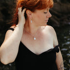 A woman with red hair wearing a delicate silver necklace with a teardrop-shaped ivory pearl pendant, matching earrings, and a pearl and silver bracelet, posing in a black off-the-swimsuit against a dark, blurred coastal background in cornwall.