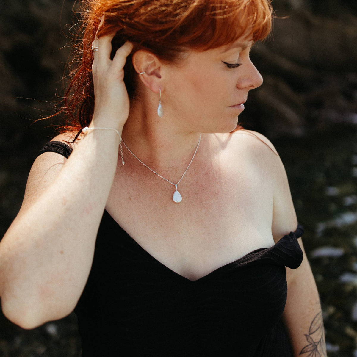 A woman with red hair wearing a delicate silver necklace with a teardrop-shaped ivory pearl pendant, matching earrings, and a pearl and silver bracelet, posing in a black off-the-swimsuit against a dark, blurred coastal background in cornwall.