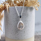 A silver necklace with a teardrop-shaped freshwater pearl pendant, displayed in front of a speckled ceramic vase filled with dried grasses.