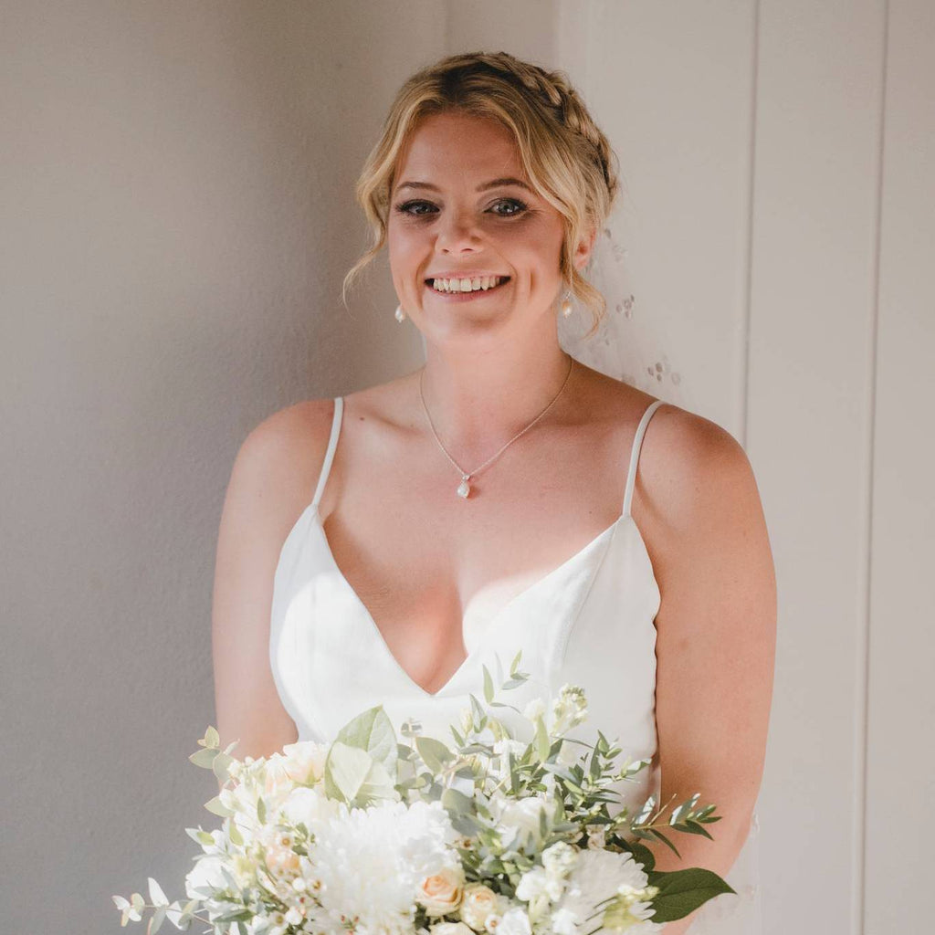 A smiling blonde bride wearing a white wedding dress with pearl jewellery set and holding a white floral bouquet against a neutral indoor background.