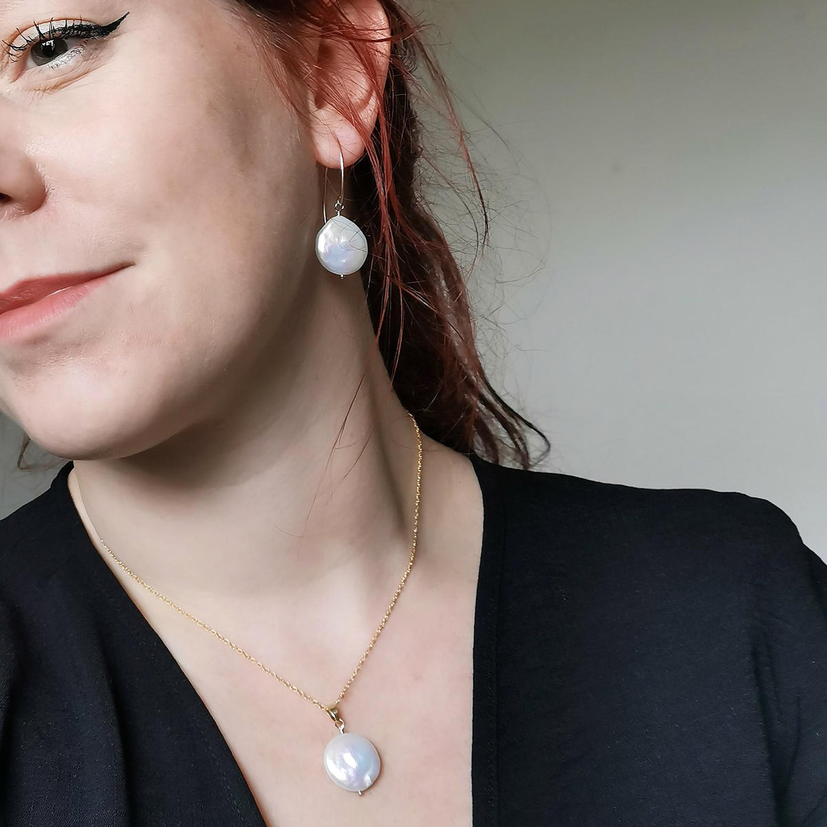 A close-up lifestyle shot of a woman wearing a gold chain necklace and matching drop earring, both featuring an ivory white freshwater coin pearl pendant, against a neutral light gray background.