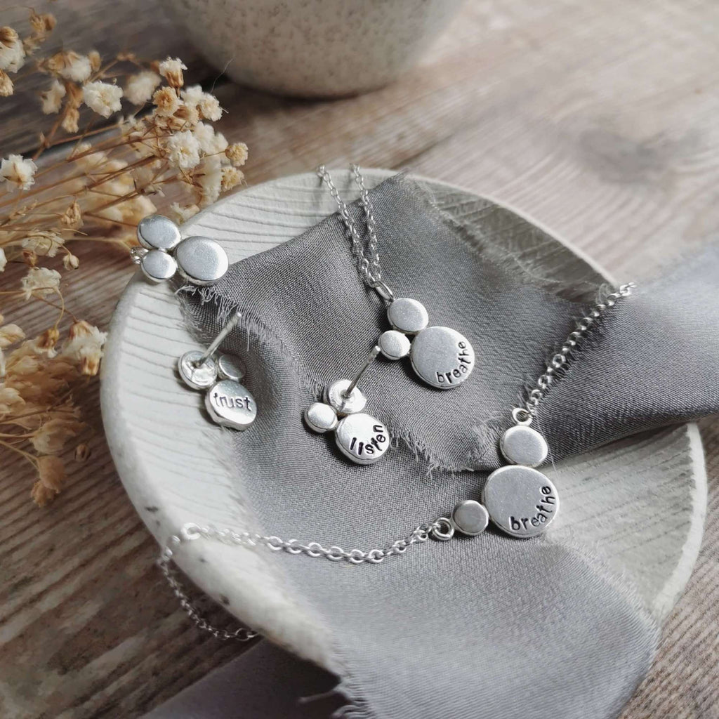 Silver necklace with clusters of silver shapes with words of affirmations stamped into them, lying on a dish with grey ribbon with dried flowers in the background