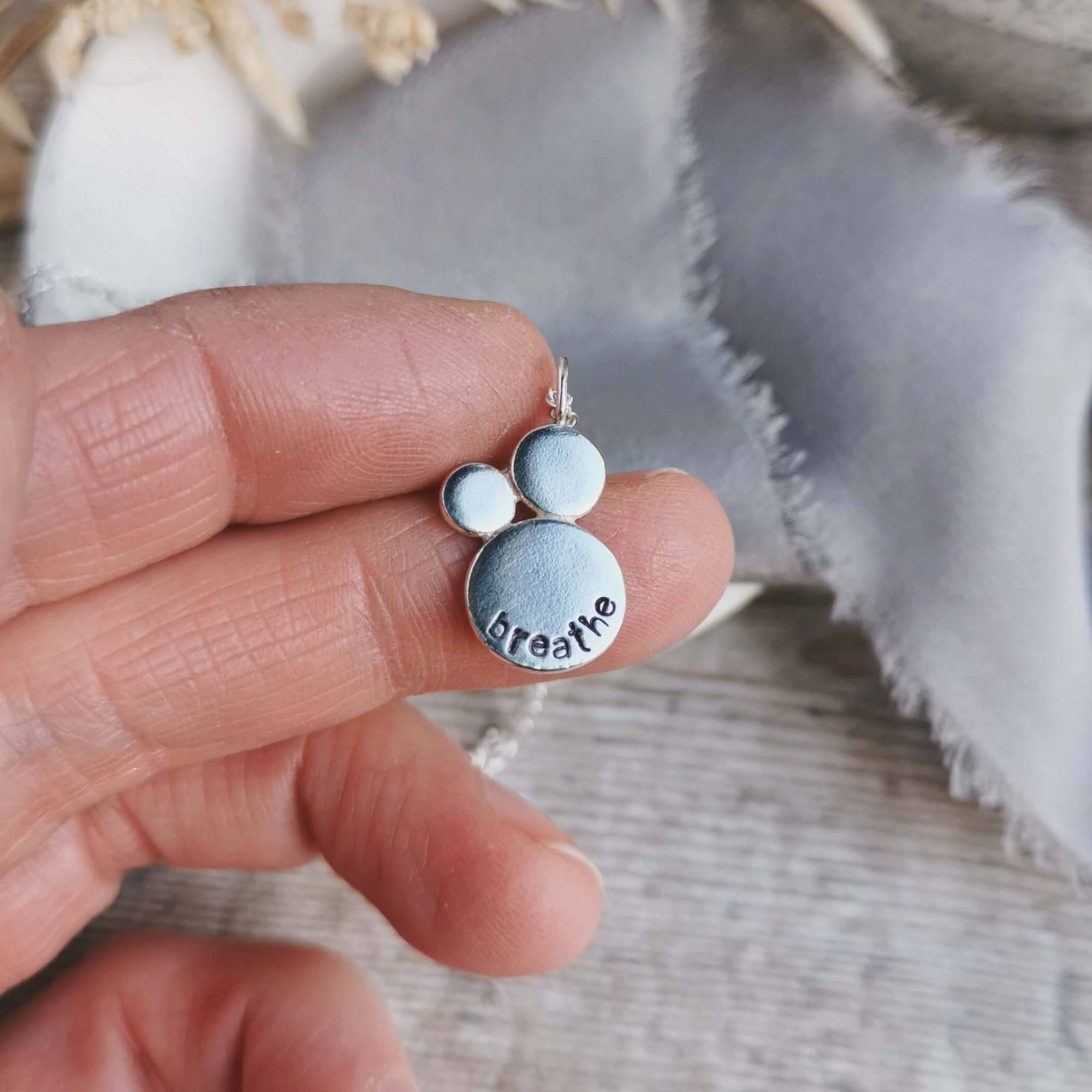 Silver pendant with 'breathe' engraving held between fingers against a textured fabric background