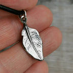 The reverse of a silver feather-shaped pendant with hallmarks, hanging from a black cord and held on fingers.