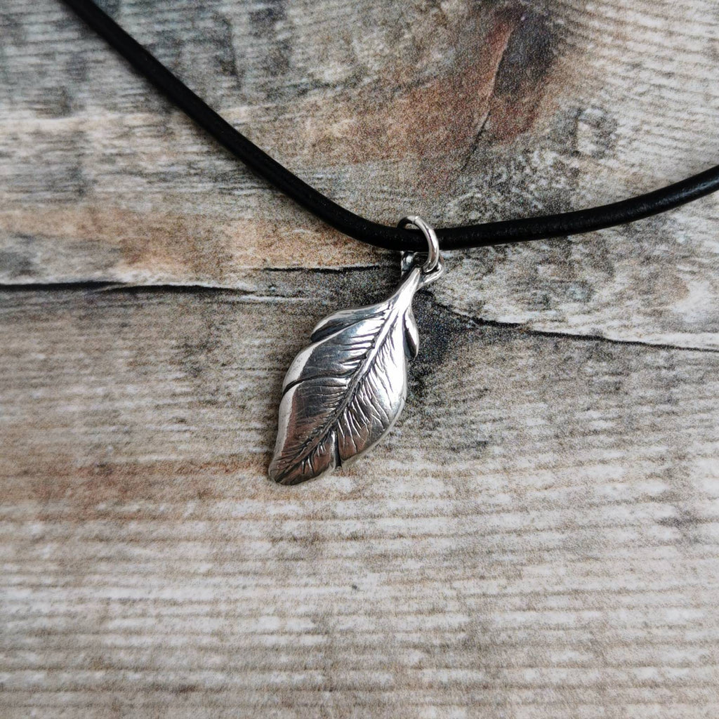 A silver feather pendant necklace on a black leather cord, displayed on a rustic weathered wood background.