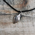 A silver feather pendant necklace on a black leather cord, displayed on a rustic weathered wood background.