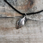 A silver feather pendant necklace on a black leather cord, displayed on a rustic weathered wood background.